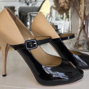 May include: A pair of high-heeled shoes with a two-tone design. The upper portion is a light tan color, while the lower part and strap are glossy black. The shoes have a peep-toe design and a thin, stiletto heel. A black buckle secures the ankle strap.