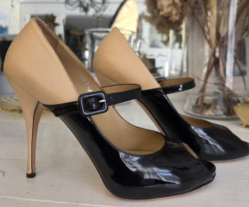 May include: A pair of high-heeled shoes with a two-tone design. The upper portion is a light tan color, while the lower part and strap are glossy black. The shoes have a peep-toe design and a thin, stiletto heel. A black buckle secures the ankle strap.