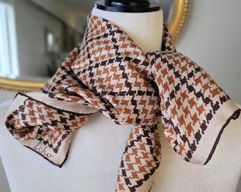 Puede incluir: Un pañuelo cuadrado con un patrón de pata de gallo en marrón y beige con un borde marrón oscuro. El pañuelo está atado alrededor del cuello de un maniquí y muestra el nombre de la marca Echo impreso en cursiva cerca de la esquina inferior.