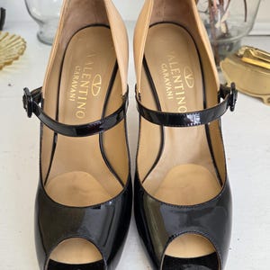 May include: Pair of black patent leather peep-toe heels with a tan interior and a black ankle strap. The heels are branded with the "Valentino Garavani" logo in gold lettering. The shoes have a classic, elegant design.