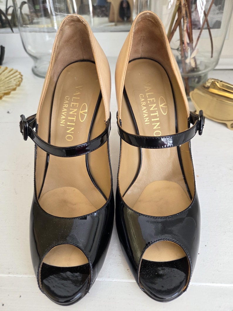 May include: Pair of black patent leather peep-toe heels with a tan interior and a black ankle strap. The heels are branded with the "Valentino Garavani" logo in gold lettering. The shoes have a classic, elegant design.