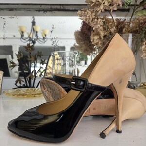 May include: A pair of high-heeled shoes with a two-tone design. The toe and strap are black patent leather, while the heel and upper are a light beige color. The shoes have a buckle closure and a stiletto heel.
