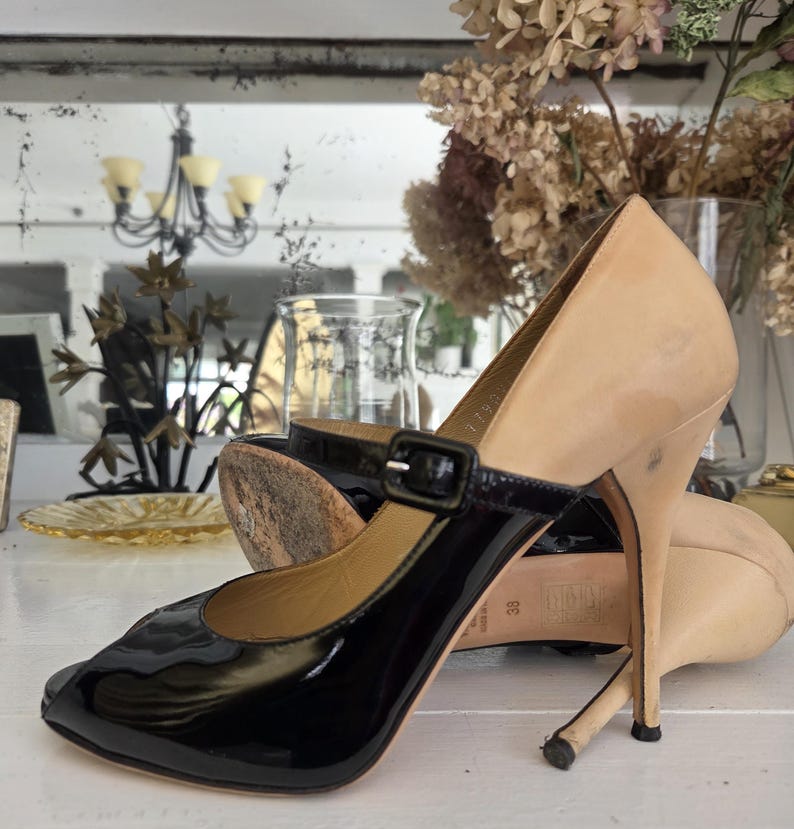 May include: A pair of high-heeled shoes with a two-tone design. The toe and strap are black patent leather, while the heel and upper are a light beige color. The shoes have a buckle closure and a stiletto heel.