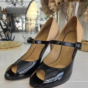 May include: A pair of black patent leather peep-toe heels with a tan upper and ankle strap. The heels have a buckle closure and a stiletto heel. The shoes are displayed on a white surface.