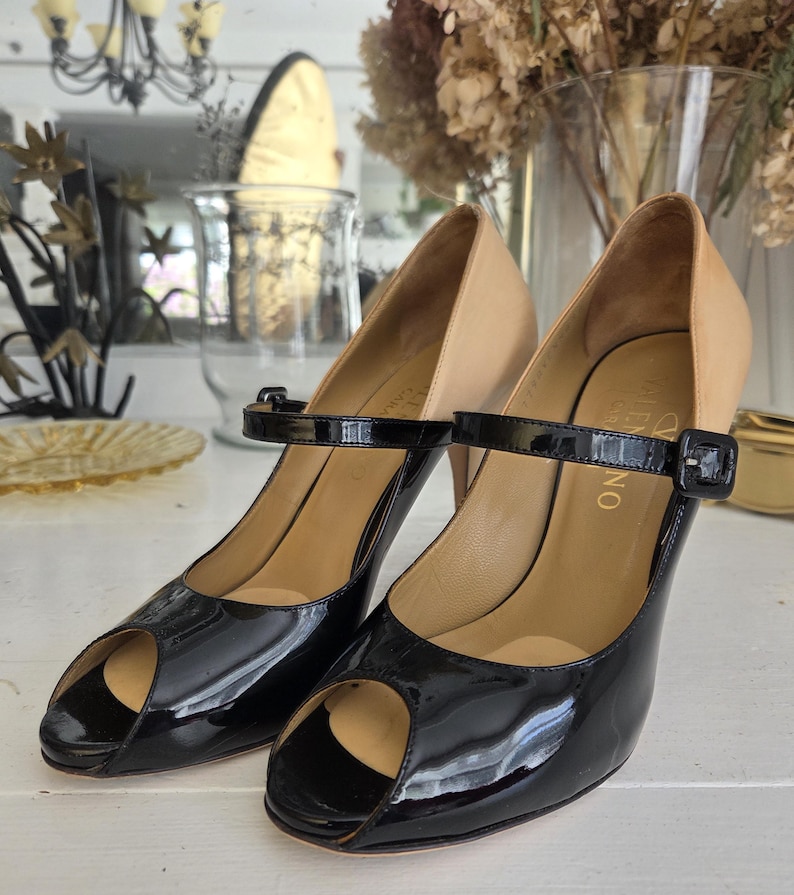 May include: A pair of black patent leather peep-toe heels with a tan upper and ankle strap. The heels have a buckle closure and a stiletto heel. The shoes are displayed on a white surface.