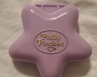 Könnte beinhalten: Ein lavendelfarbener Polly Pocket-Kompakt in Sternform. Der Kompakt hat ein goldfarbenes ovales Emblem mit den Worten "Polly Pocket" und einem Herzsymbol. Der Kompakt ist geschlossen und hat ein kleines, erhöhtes Scharnier.