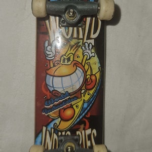 May include: A miniature skateboard with a colorful graphic design. The deck features a cartoon character with a smiling face and the words "World Industries" and "Industries" in white. The wheels are off-white.