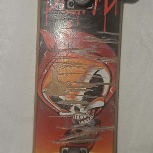 Pode incluir: Um pequeno fingerboard de madeira com um gráfico vermelho, laranja e preto de uma caveira usando um chapéu. O fingerboard tem rodas brancas e trucks pretos.