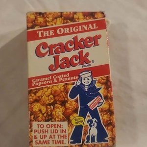 May include: A red, white, and blue box of Cracker Jack caramel coated popcorn and peanuts. The box features a cartoon sailor holding a box of Cracker Jack and a dog. The text on the box reads "The Original Cracker Jack Brand" and "Caramel Coated Popcorn & Peanuts".
