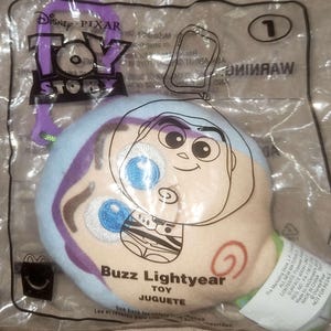 May include: A Buzz Lightyear plush toy from the Disney Pixar Toy Story franchise, packaged in a clear plastic bag. The toy features an embroidered face and the text "Buzz Lightyear TOY JUGUETE". A purple Disney Pixar Toy Story keychain is attached.