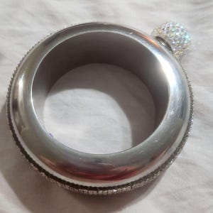 May include: A silver-coloured, circular flask with a crystal-studded cap. The flask's outer rim is also adorned with small, sparkling crystals. The design is sleek and modern, suitable for carrying beverages.