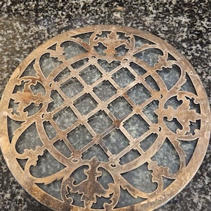 May include: A circular, silver-toned metal coaster with a decorative, intricate design. The design features a repeating pattern of squares and floral motifs.