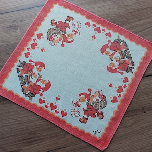 Könnte beinhalten: Ein rotes und weißes Baumwolltaschentuch mit einem festlichen Weihnachtsmotiv mit kleinen Wichteln, die Geschenke und Herzen tragen. Das Taschentuch hat einen gewellten Rand und die Initialen "IM" in der rechten unteren Ecke.