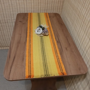 以下が含まれることがあります： 木製のテーブルの上に、フリンジの付いた黄色とオレンジのストライプのテーブルランナーが置かれています。ランナーの上に小さな装飾品が置かれています。