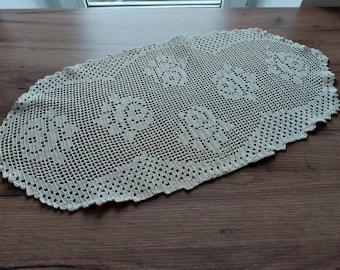 May include: Cream-colored, oval-shaped crocheted doily with a floral pattern. The openwork design features repeating floral motifs and a scalloped edge. The doily is placed on a dark wooden surface.