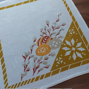 Op de afbeelding: Wit linnen placemat met een gele rand en een ontwerp van paaseieren en wilgentakken in de hoek.