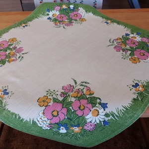 Könnte beinhalten: Eine beige Tischdecke mit grünem Rand und Blumendruck. Die Blumen sind rosa, gelb und blau. Die Tischdecke ist rechteckig und hat einen Fransenrand.