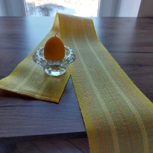 May include: A yellow and green striped table runner with a yellow candle in a glass holder.