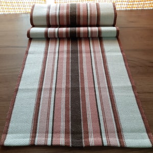 Op de afbeelding: Een gestreepte tafelrunner in tinten van bruin, roze en wit. De runner is in drieën gevouwen en ligt op een houten oppervlak.