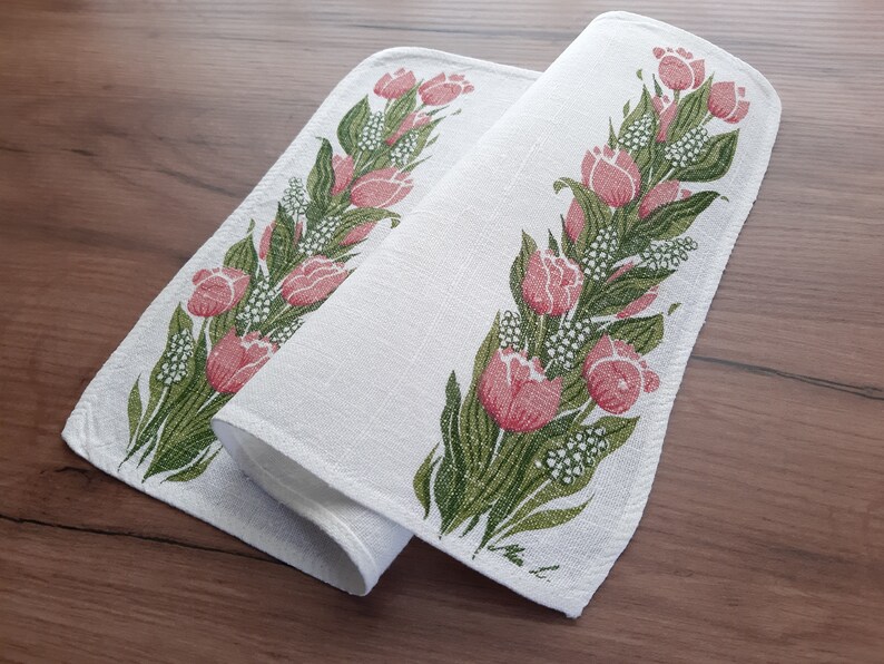 May include: Two white linen tea towels with a pink and green floral design. The towels feature a repeating pattern of tulips and leaves.