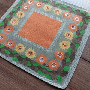 Può includere: Un tovaglietta quadrata in tessuto con un motivo arancione e verde e un bordo marrone. La tovaglietta ha un design floreale con un quadrato arancione centrale.