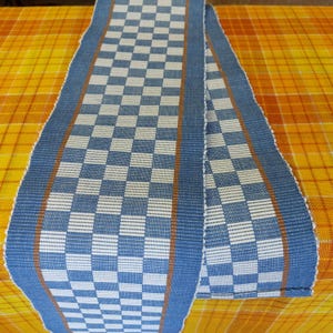 Puede incluir: Un camino de mesa a cuadros azules y blancos con ribete naranja. El camino de mesa está doblado por la mitad y está sobre una superficie a cuadros amarilla y naranja.