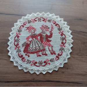 May include: Round, cream-colored doily with a detailed embroidered design. The center features two figures in traditional attire, rendered in red and brown. The doily is edged with a delicate, scalloped lace trim.