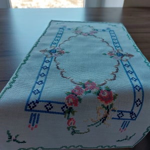 May include: A white table runner with a blue and green embroidered border and floral design. The floral design features pink, red, and yellow flowers.