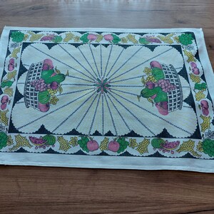 May include: A white cotton tea towel with a black border and a colorful fruit design. The design features two baskets filled with grapes, apples, and cucumbers. The baskets are surrounded by a repeating pattern of fruit and leaves.