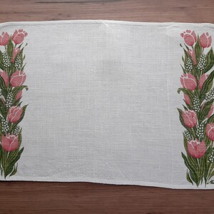 May include: White linen placemat with a floral design featuring pink tulips and green leaves. The placemat has a subtle texture and a slightly frayed edge.