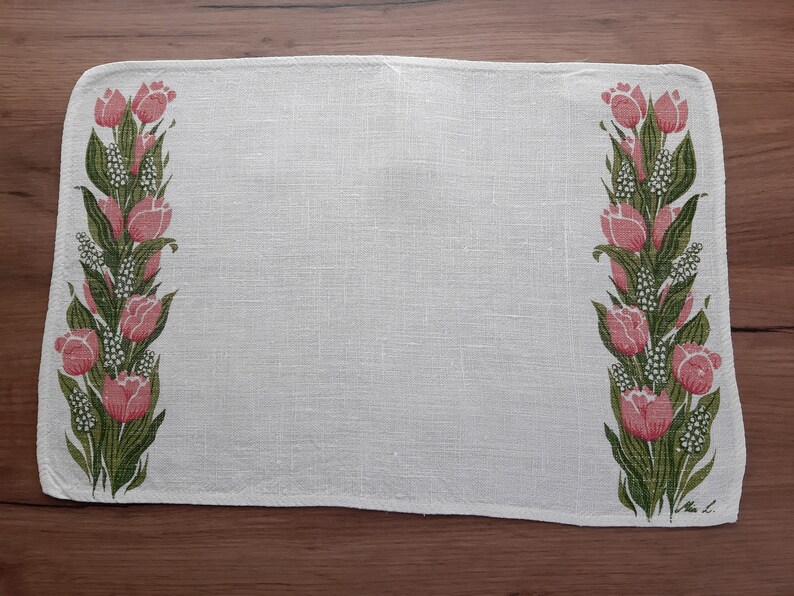 May include: White linen placemat with a floral design featuring pink tulips and green leaves. The placemat has a subtle texture and a slightly frayed edge.