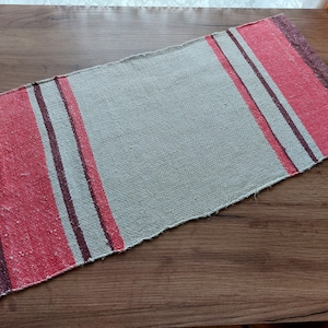 May include: A woven rug with a light gray background and stripes of red and brown. The rug is made of a coarse material and has a rustic look.