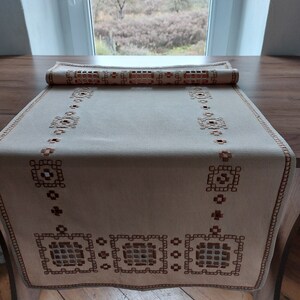 Puede incluir: Un camino de mesa de lino beige con diseños geométricos bordados en marrón. El camino de mesa tiene un borde festoneado y está doblado por la mitad.