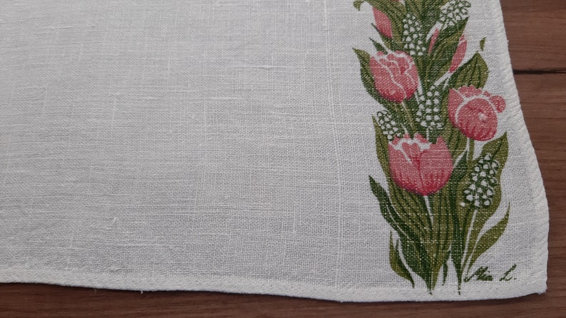 May include: White linen tea towel with a green and pink floral design. The design features tulips and small white flowers. The towel is signed "Mia L."