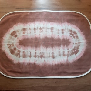 Könnte beinhalten: Ein brauner, ovaler Stoff mit Batik-Muster in Weiß und Rosa. Der Stoff hat einen weißen Rand.