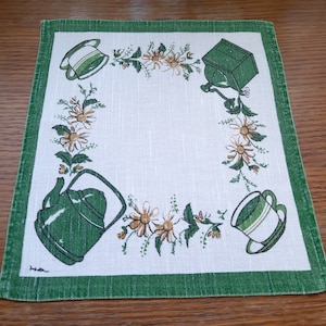 Könnte beinhalten: Ein weißes Leinen-Geschirrtuch mit grünem Rand. Das Tuch zeigt ein grünes Blumenmuster mit Teekannen, Kaffeemühlen und Tassen.
