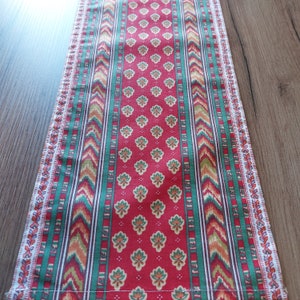 Puede incluir: Un camino de mesa rojo, verde y blanco con un patrón floral. El camino de mesa tiene un patrón geométrico en los lados.