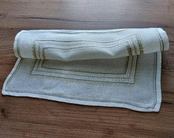 May include: A white linen table runner with a yellow and blue embroidered border. The runner is folded in half and lying on a wooden surface.
