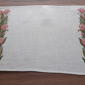 May include: White linen placemat with a pink and green floral design featuring tulips. The placemat has a slightly scalloped edge.