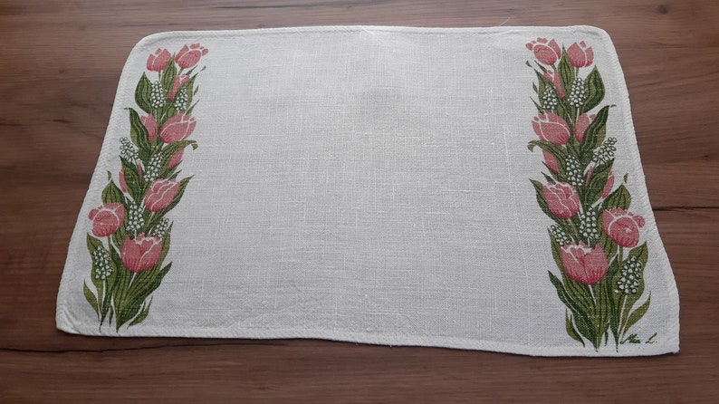 May include: White linen placemat with a pink and green floral design featuring tulips. The placemat has a slightly scalloped edge.