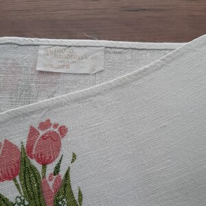 May include: White linen tea towel with a pink and green floral design featuring tulips. The towel has a label that reads "Stero Handtryck AB 100% Linne Made in Sweden".