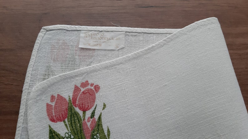 May include: White linen tea towel with a pink and green floral design featuring tulips. The towel has a label that reads "Stero Handtryck AB 100% Linne Made in Sweden".