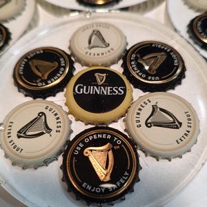 May include: A collection of Guinness bottle caps arranged on a clear plate. The caps feature the Guinness logo, a harp, and text such as "Enjoy Safely" and "Extra Stout". The caps are black, gold, and white.