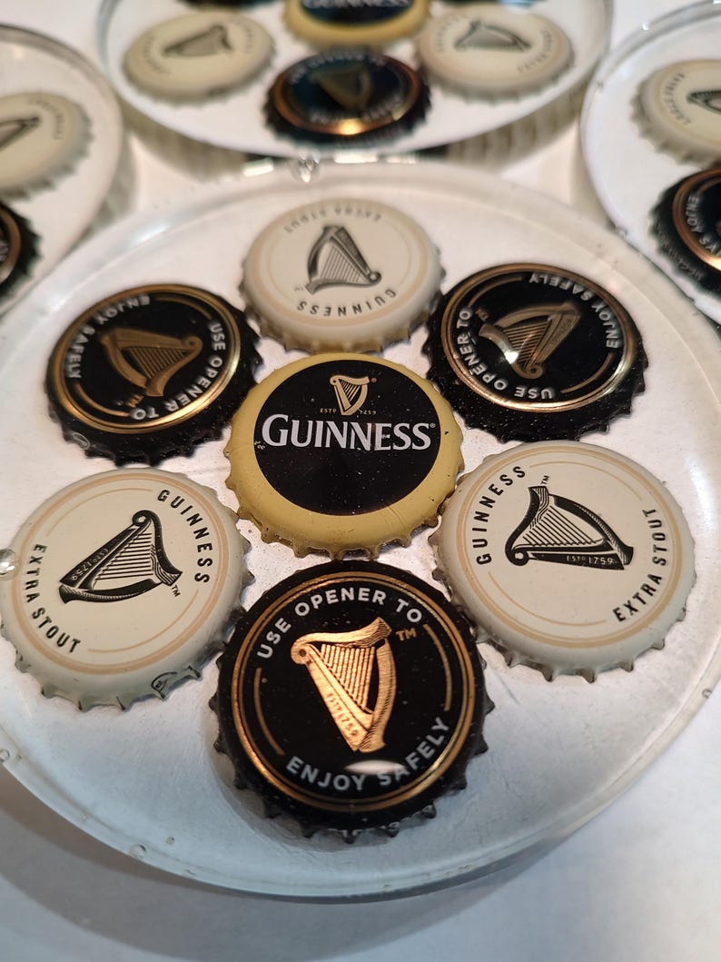 May include: A collection of Guinness bottle caps arranged on a clear plate. The caps feature the Guinness logo, a harp, and text such as "Enjoy Safely" and "Extra Stout". The caps are black, gold, and white.