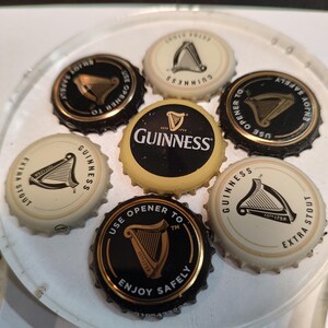 May include: A collection of Guinness bottle caps, featuring the iconic harp logo. The caps are black and gold, and white and gold, with text that reads "Guinness", "Extra Stout", and "Enjoy Safely".