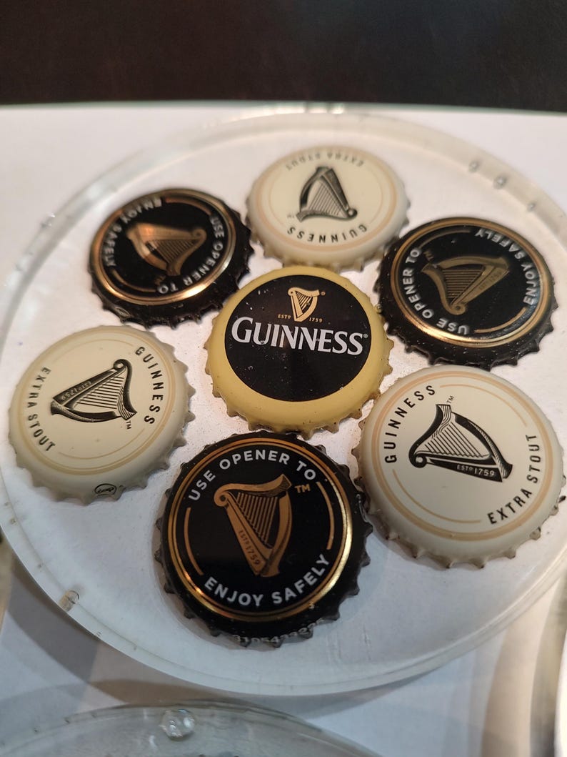 May include: A collection of Guinness bottle caps, featuring the iconic harp logo. The caps are black and gold, and white and gold, with text that reads "Guinness", "Extra Stout", and "Enjoy Safely".
