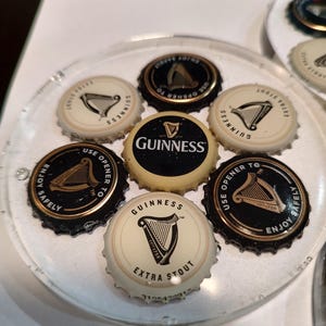 May include: A clear, circular coaster featuring several Guinness bottle caps. The caps are black and gold, with the Guinness logo and text. Some caps read "Extra Stout" and "Use Opener To Enjoy Safely."