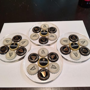 May include: Four clear resin coasters, each embedded with beer bottle caps. The caps feature the Guinness logo and other designs. The coasters are arranged on a white surface, showcasing a set of drink accessories.