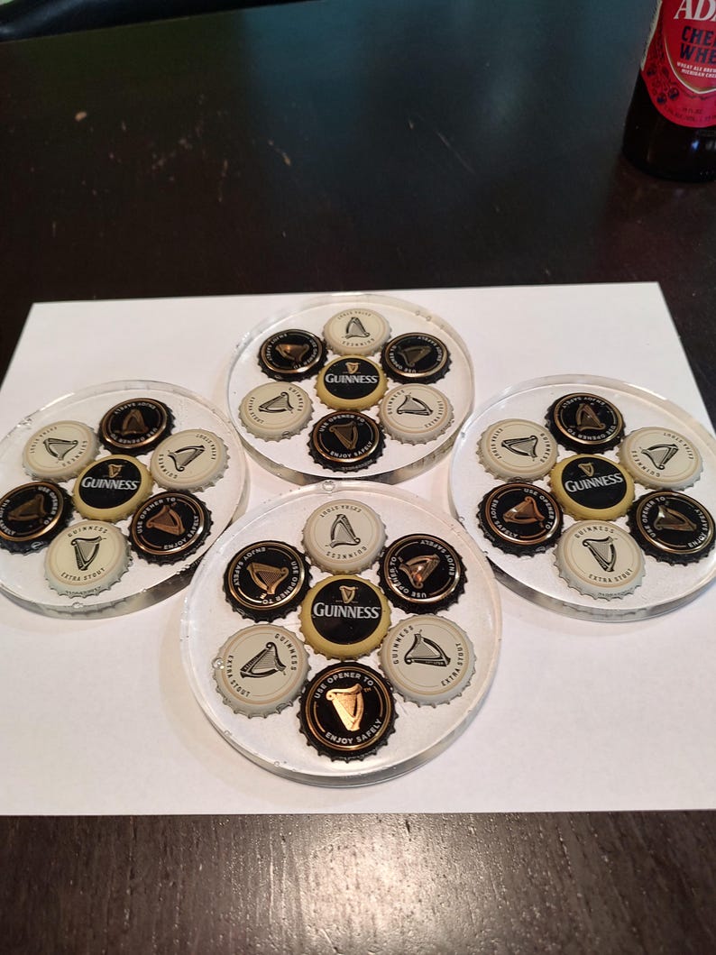 May include: Four clear resin coasters, each embedded with beer bottle caps. The caps feature the Guinness logo and other designs. The coasters are arranged on a white surface, showcasing a set of drink accessories.