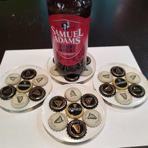 May include: A bottle of Samuel Adams Cherry Wheat beer rests on a clear coaster surrounded by other coasters. Each coaster is decorated with beer bottle caps, including Guinness caps. The coasters are on a dark surface.